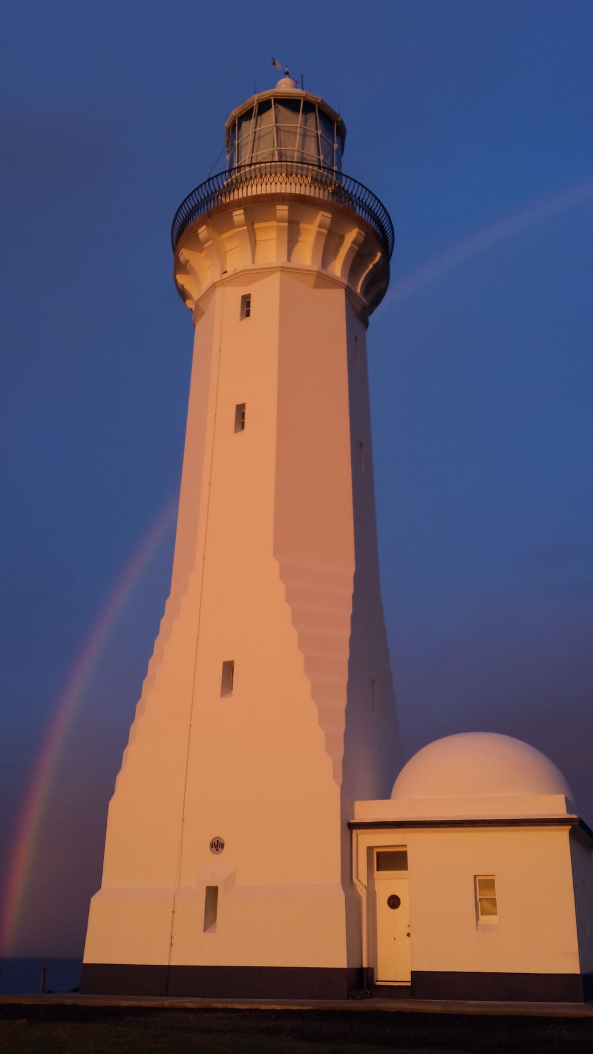 Green Cape Lighthouse lit warm at sunrise against a deep blue sky, with part of a rainbow behind it