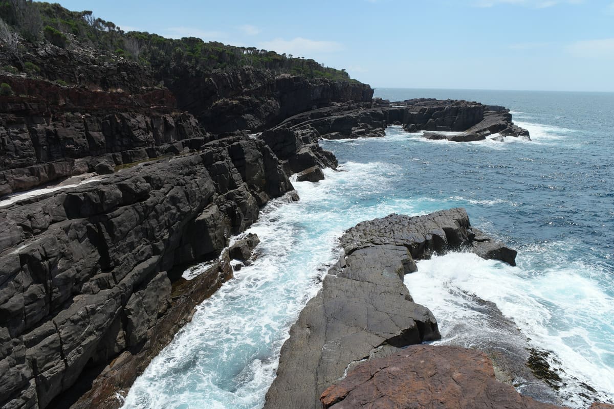 Dark layered rock cliffs and ledges meeting breaking surf, with a forested headland beyond