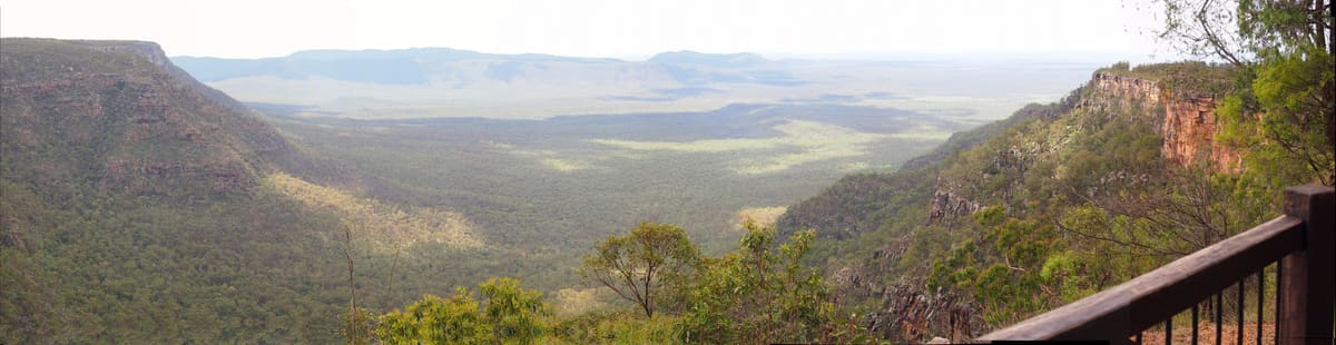 Panorama from a lookout over a sunlit valley and red cliff walls, timber railing at lower right