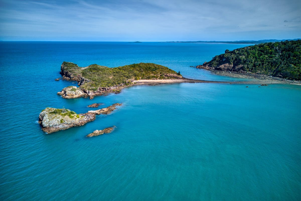 Aerial view of a turquoise bay, rocky islet and green forested headland on a sunny day