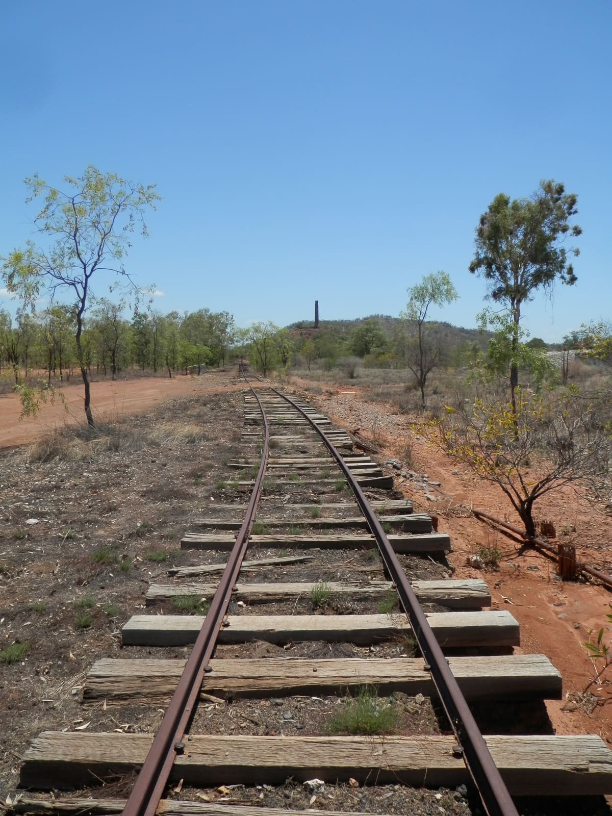Rusty disused railway tracks curving into dry red-earth bushland under clear blue sky