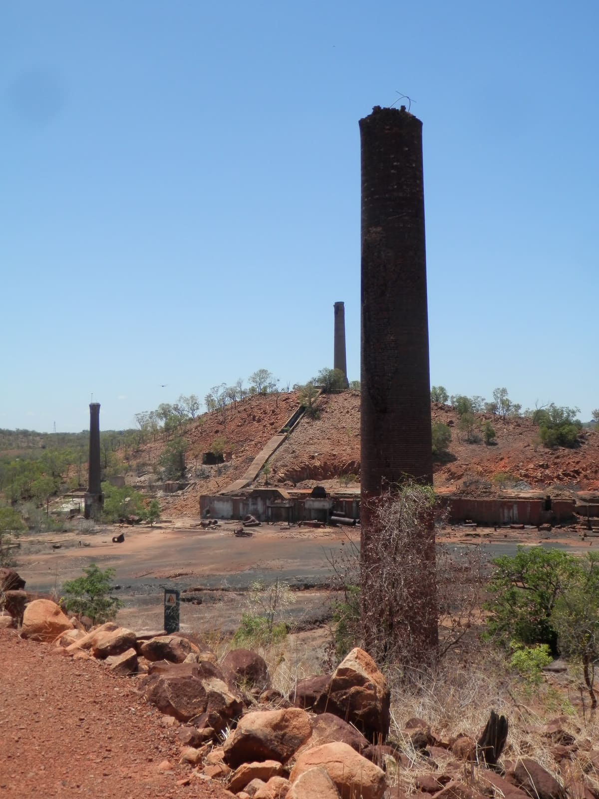 Tall dark smelter chimney close-up against red rubble and ruined hillside