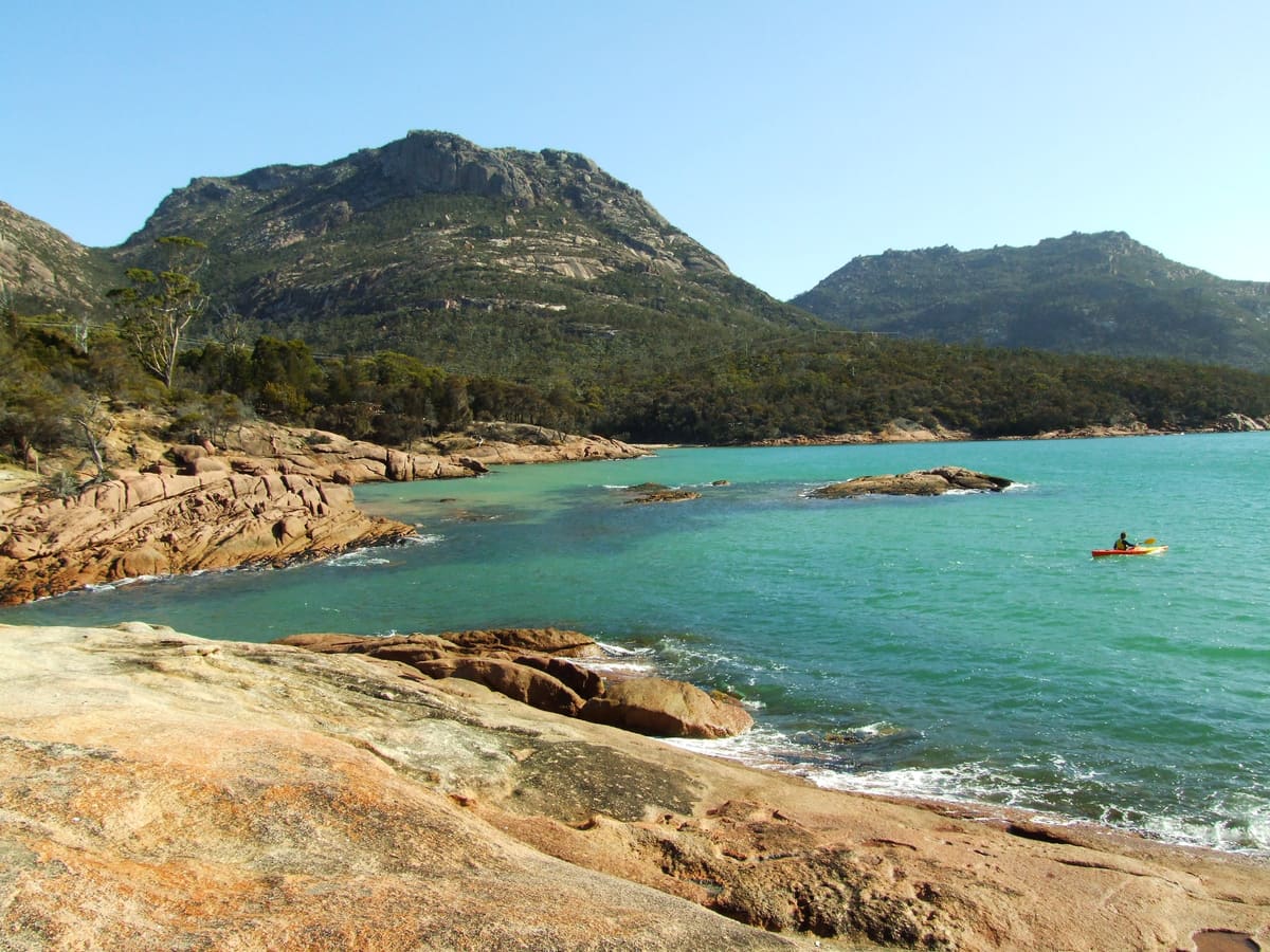 A kayaker on turquoise water in Honeymoon Bay, granite shore in front and the Hazards behind