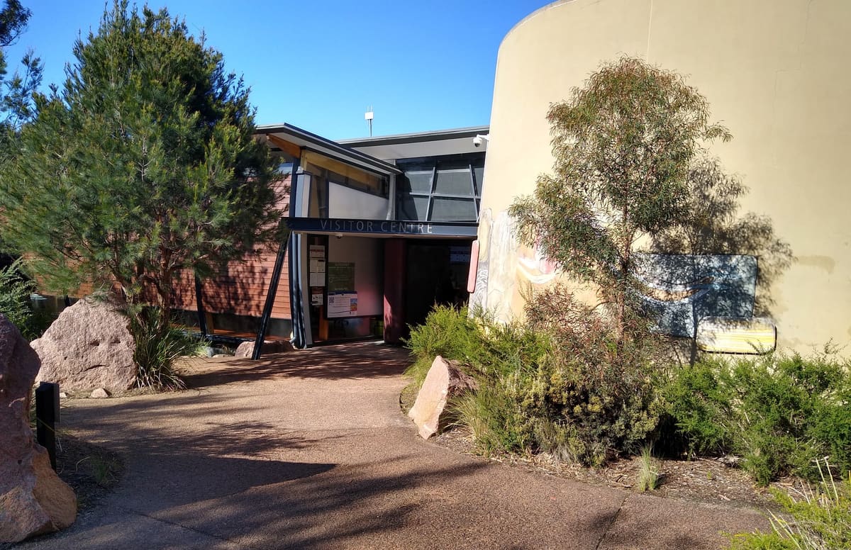 Freycinet National Park visitor centre building with curved wall, gravel path, trees and shrubs in sunlight