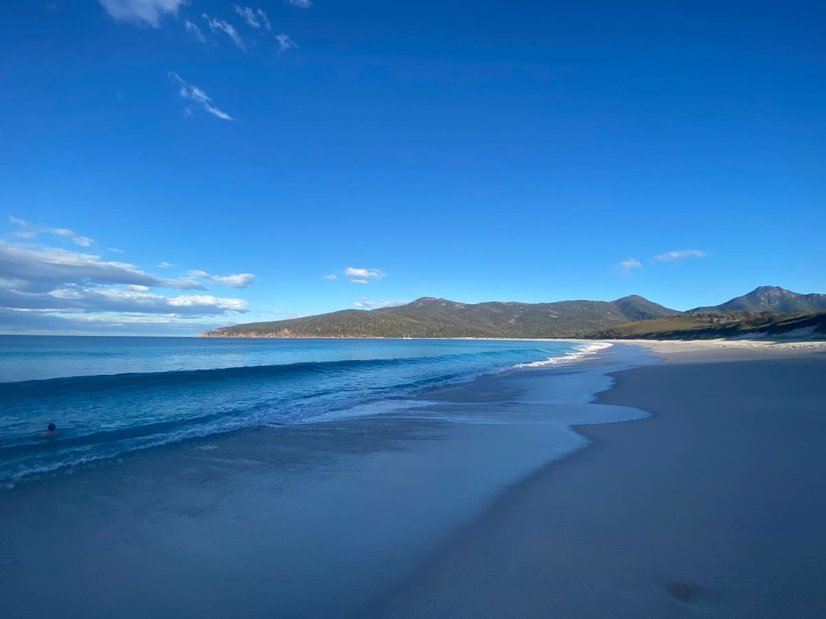 Wineglass Bay beach at the water's edge, low waves on the sand and forested hills along the far shore under a deep blue sky