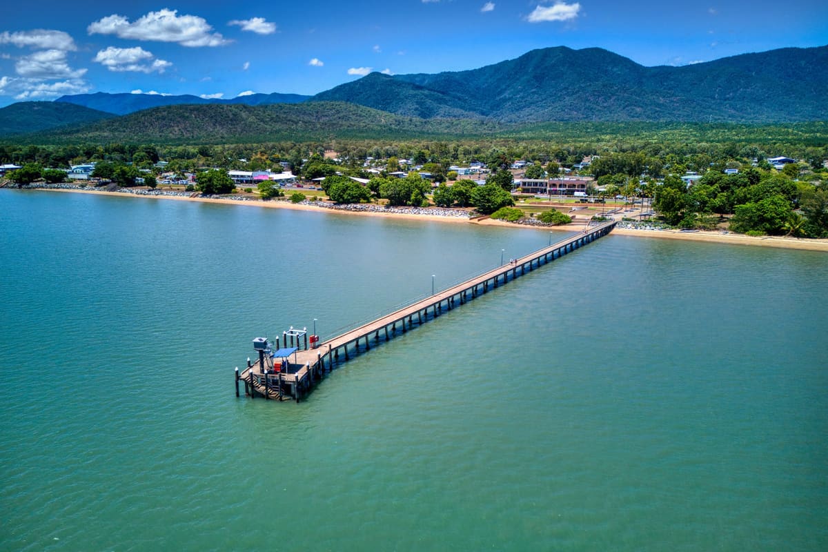 Aerial view of a long jetty over blue water, with a township and an island range behind