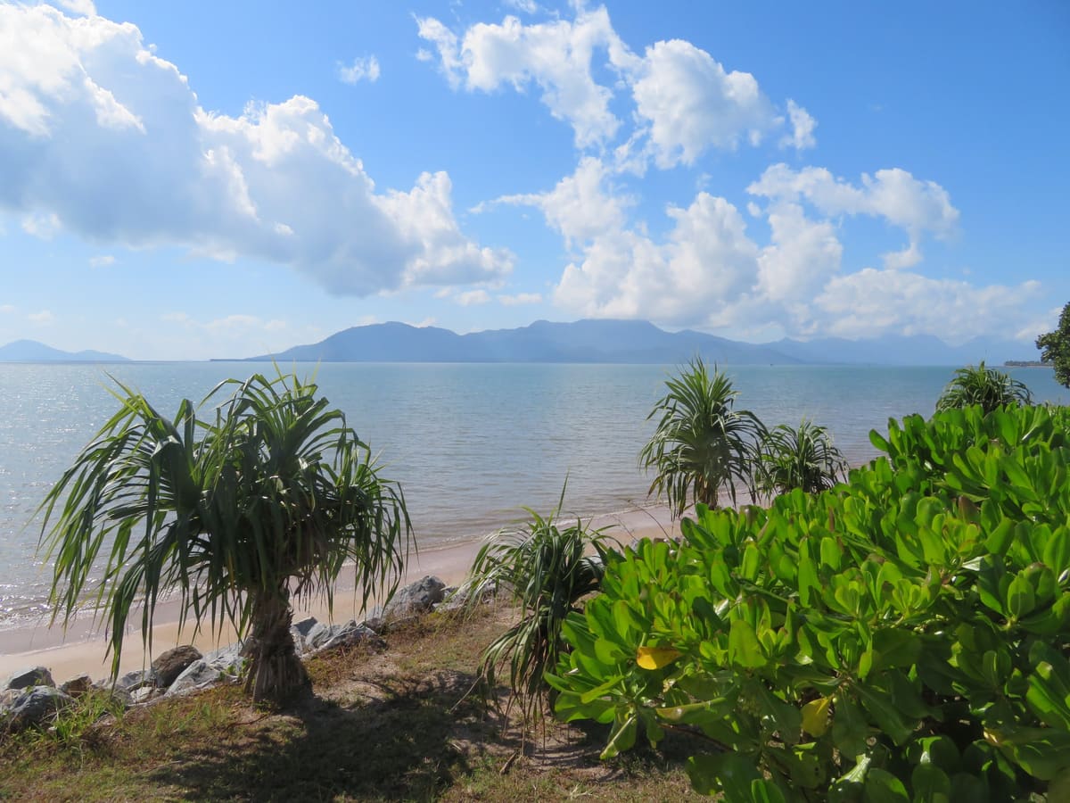 Pandanus-fringed beach, calm blue water and a mountain range across the channel beneath cumulus clouds