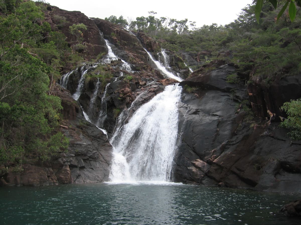 Multi-tiered white waterfall cascading down dark slabs into a green rainforest pool