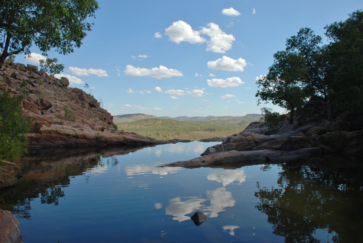 A cliff-top pool reflecting clouds, with rocks and trees framing a green plain beyond