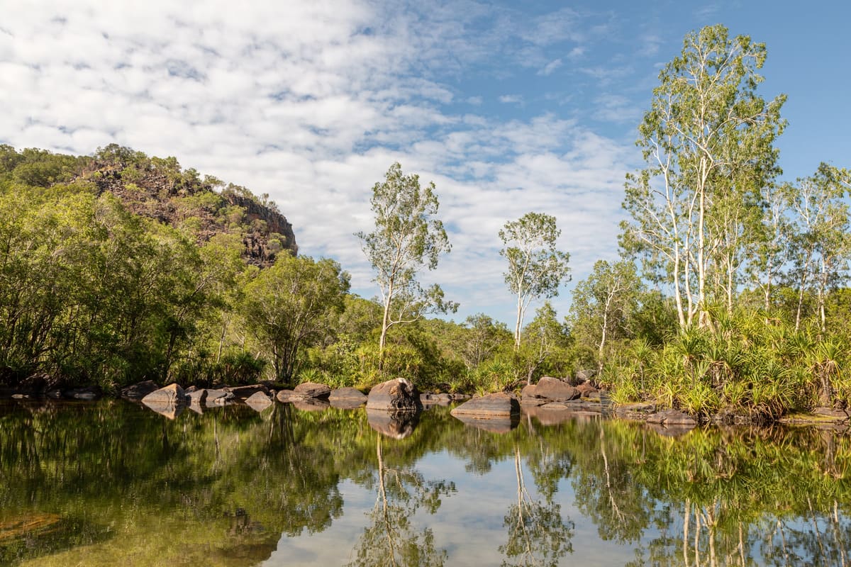 A creek pool with boulders and paperbarks below an escarpment, blue sky with clouds
