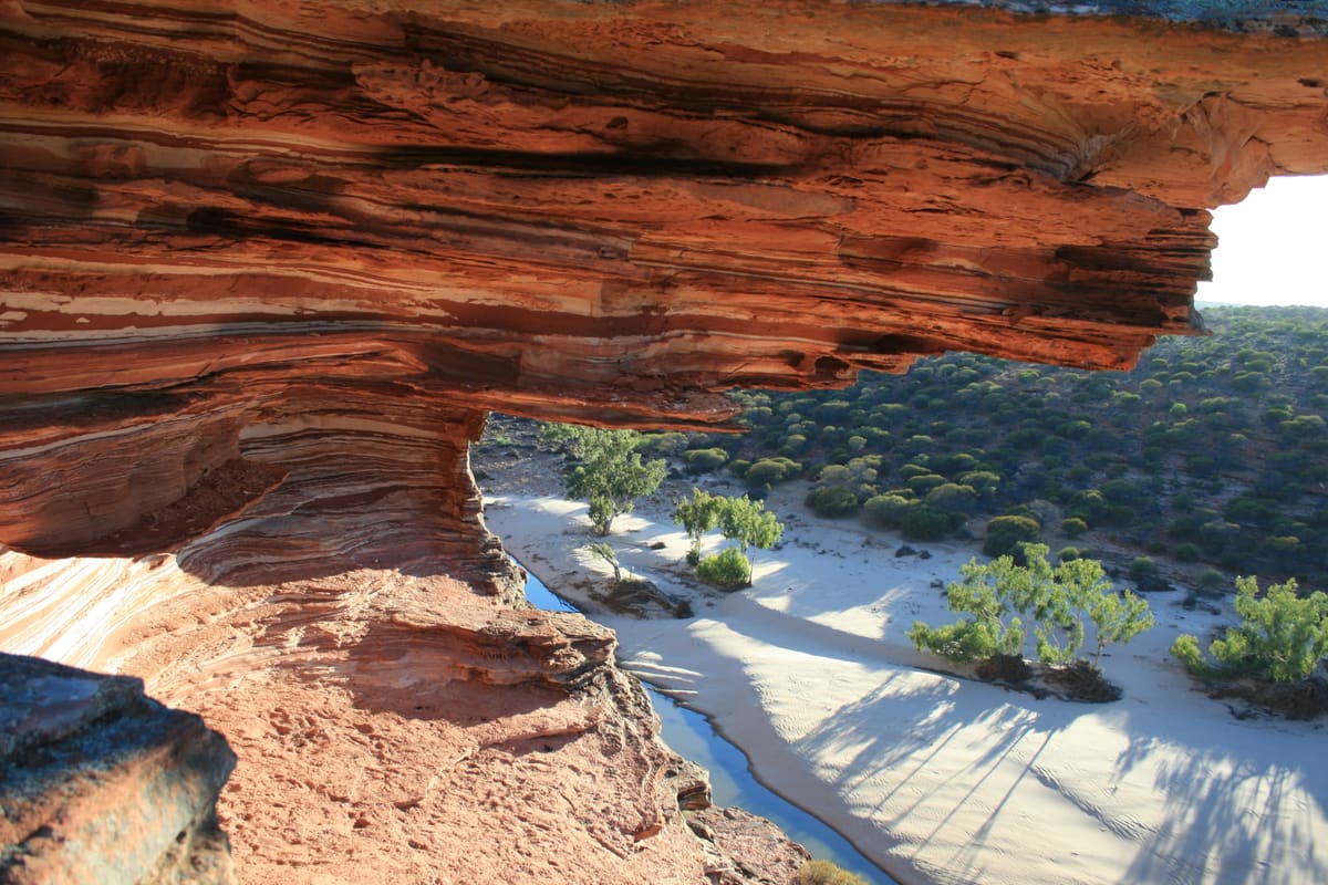 An overhanging red rock ledge framing the sandy bed of the Murchison River and trees below