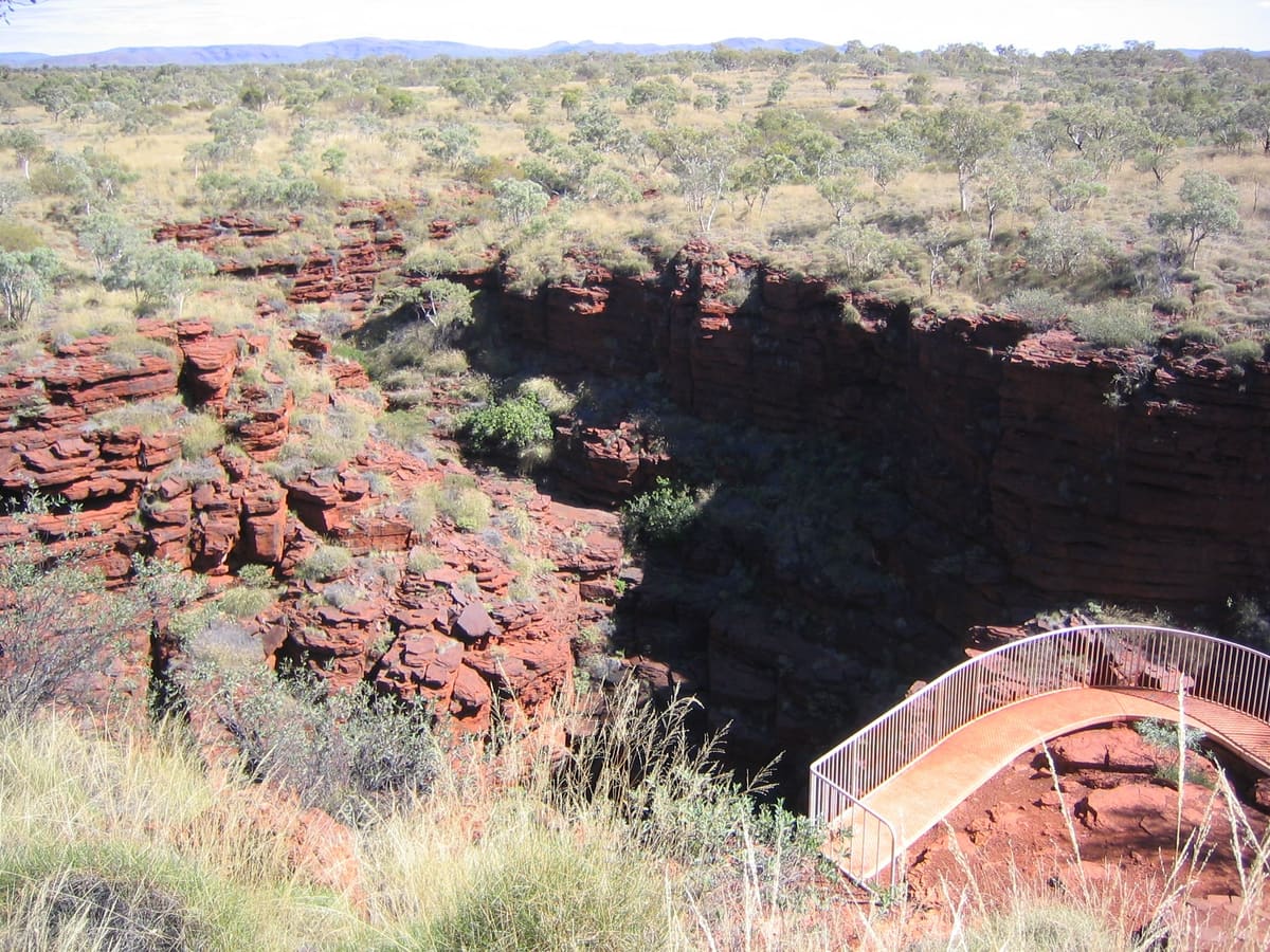A curved metal-railed walkway along the rim of a Karijini gorge, red rock walls below