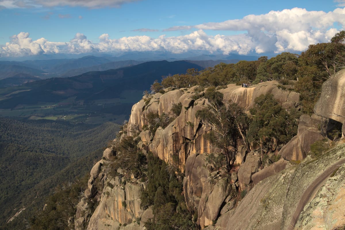 Golden granite cliff outcrop above forested valleys and distant blue ranges under cumulus clouds