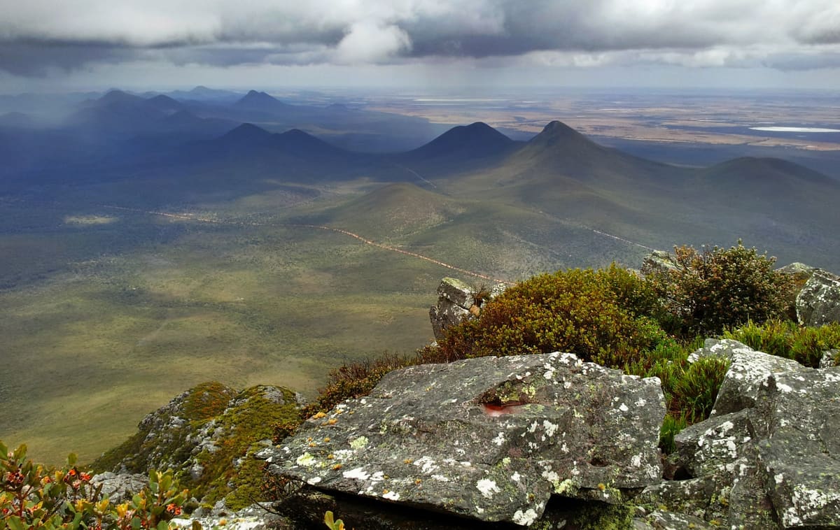 Summit panorama over jagged ranges and plains, lichen-covered rocks in foreground