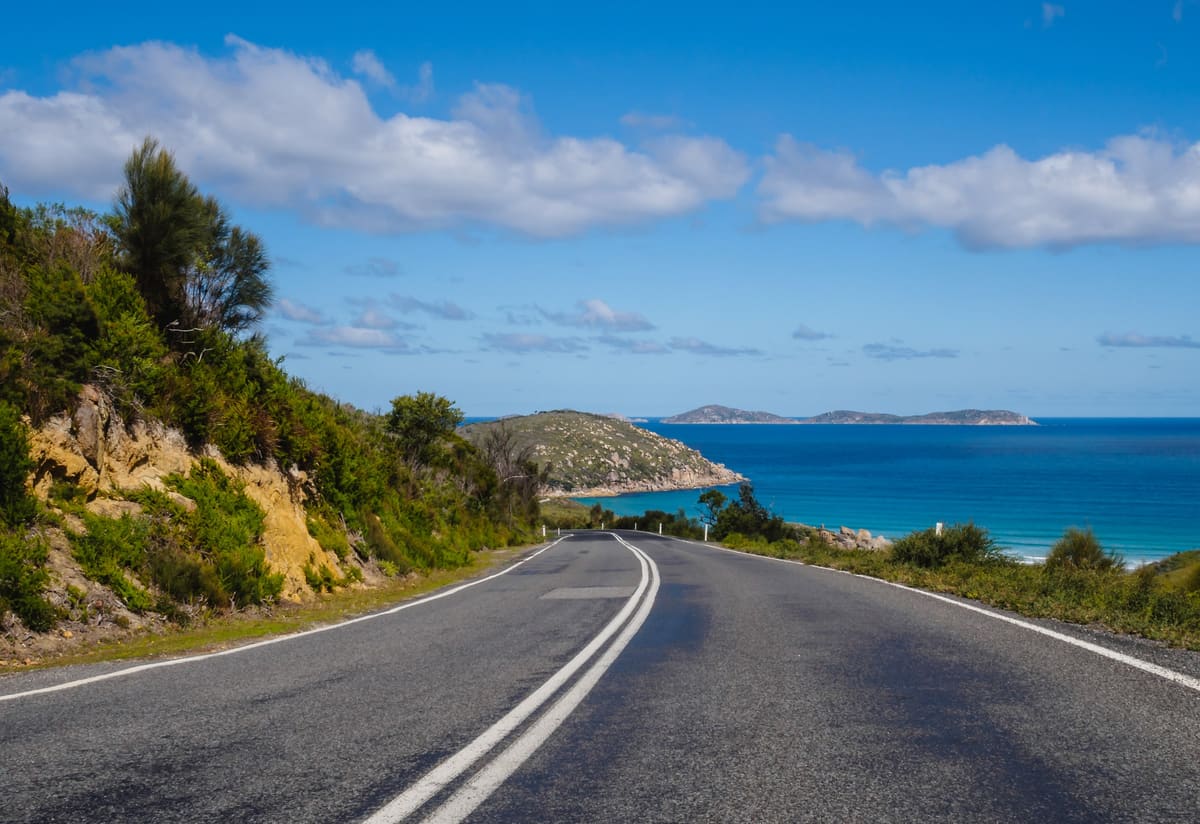 Coastal road curving toward a turquoise bay with islands and headlands under a blue sky